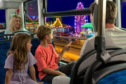 Eine Familie sitzt im Bus, der Junge zeigt aus dem Fenster, alle schauen raus und sehen einen Jahrmarkt